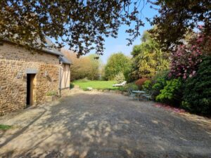 Cour en gravier devant le gîte en pierre, ouverte sur la pelouse et les massifs du jardin